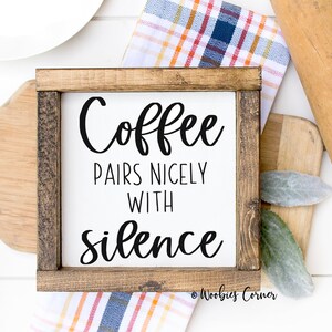 May include: A wooden sign with a white background and black text that reads "Coffee pairs nicely with silence". The sign is framed in a dark brown wood frame. The sign is sitting on a white surface with a wooden rolling pin and a white napkin.