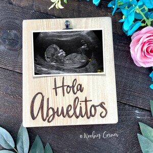 May include: A wooden picture frame with a clip at the top. The frame has a black and white ultrasound image inside and the words "Hola Abuelitos" engraved on the front. The frame is sitting on a wooden surface.