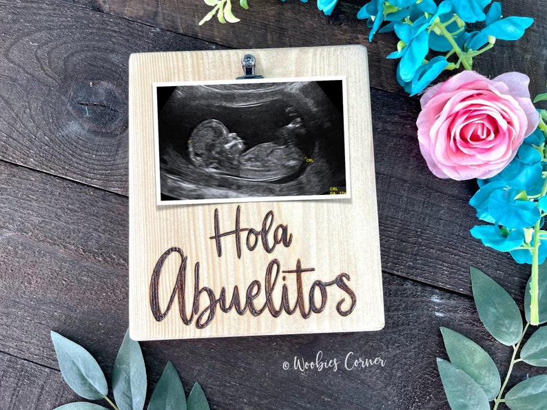 May include: A wooden picture frame with a clip at the top. The frame has a black and white ultrasound image inside and the words "Hola Abuelitos" engraved on the front. The frame is sitting on a wooden surface.