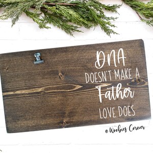 May include: A dark brown wooden sign with a metal clip at the top. The sign has white text that reads "DNA doesn't make a father, love does." The sign is on a white brick background with green foliage.