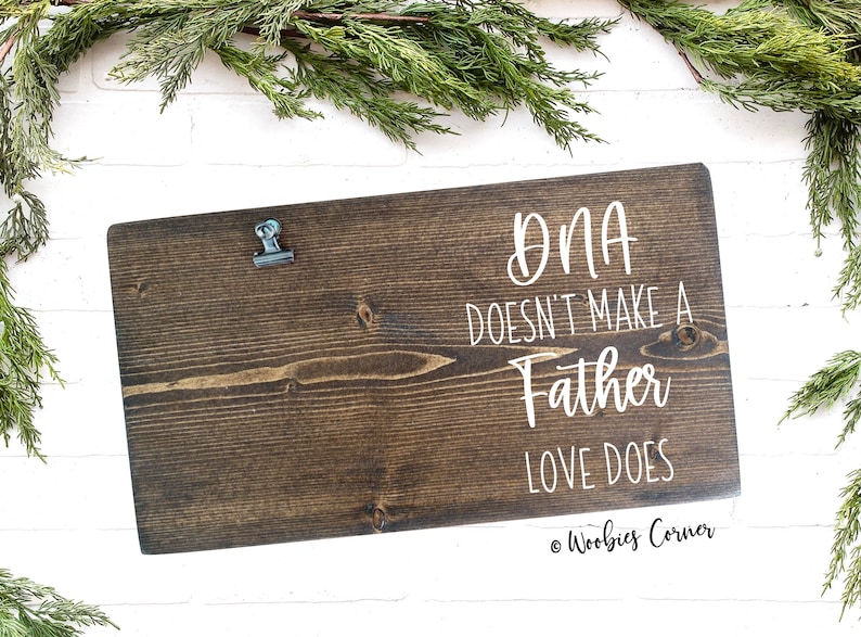 May include: A dark brown wooden sign with a metal clip at the top. The sign has white text that reads "DNA doesn't make a father, love does." The sign is on a white brick background with green foliage.