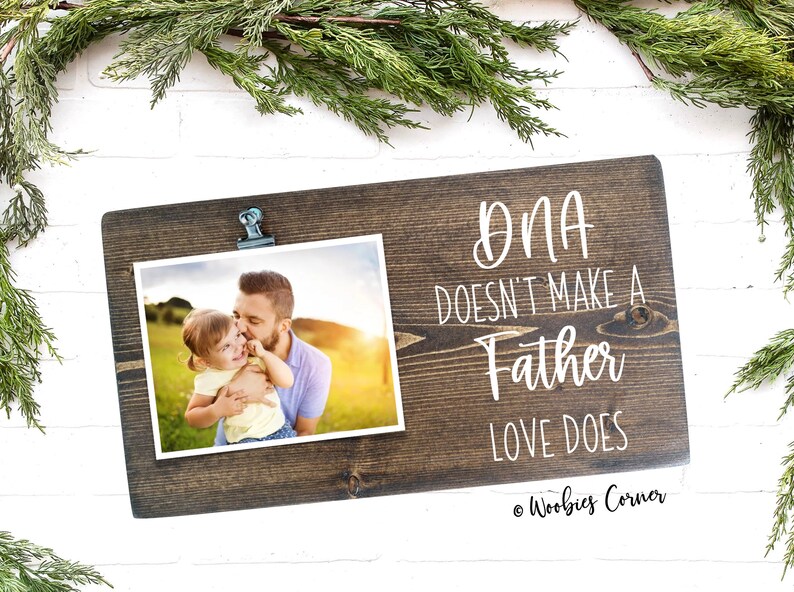 May include: A rustic wooden photo frame with a clip to hold a photo. The frame has the text "DNA doesn't make a father, love does" written on it. The photo shows a father and daughter in a field.