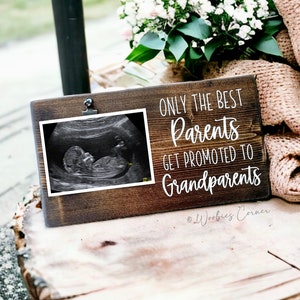 May include: A wooden sign with a white background and black text that reads "Only the best parents get promoted to grandparents". There is a photo clip on the sign and a black and white ultrasound image is in the clip. The sign is on a wooden surface.