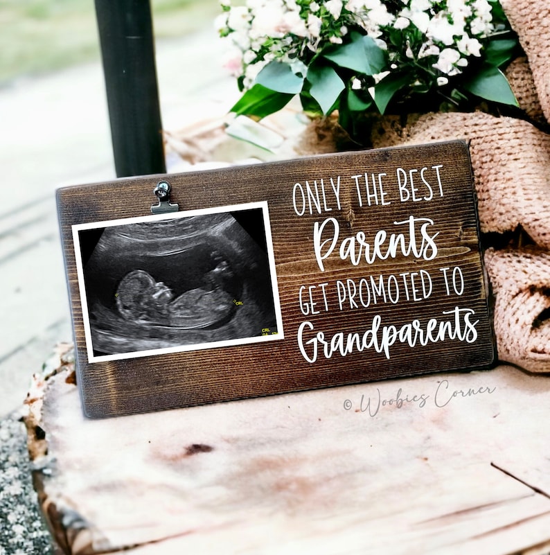 May include: A wooden sign with a white background and black text that reads "Only the best parents get promoted to grandparents". There is a photo clip on the sign and a black and white ultrasound image is in the clip. The sign is on a wooden surface.