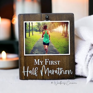 May include: A wooden photo frame with a clip at the top. The frame has a photo of a person running on a path. The text "My First Half Marathon" is written on the frame. The text "Woobies Corner" is written in the bottom right corner of the frame.