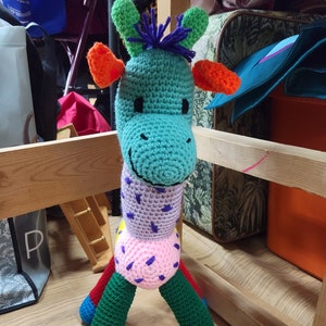 May include: A colorful crocheted giraffe toy with a green head, purple and pink body, and blue and green legs. The giraffe has orange ears and a purple tuft of hair on its head.
