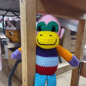 May include: A colorful crocheted monkey toy with a pink head, green eyes, yellow smile, purple, red, blue, and orange stripes, and yellow feet.