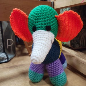 May include: A crocheted elephant toy with a green head, orange ears, a white trunk, and a multi-colored body. The body is made of purple, green, blue, and yellow yarn.