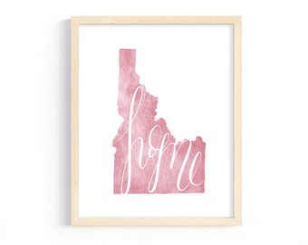 May include: Framed art print featuring a watercolor-style map of Idaho in a dusty rose hue. The word "home" is written in elegant white script across the map. The print is set in a light wood frame.