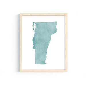 May include: A framed print featuring a teal watercolor map of Vermont. The map is centered on a white background, enclosed within a light wood-colored frame. The artwork has a minimalist aesthetic.
