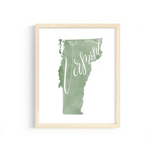 May include: A framed print featuring a watercolor-style map of Vermont in shades of green. The word "Vermont" is written in elegant white script across the map. The frame is a light wood color.