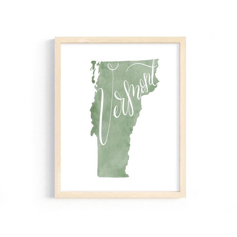 May include: A framed print featuring a watercolor-style map of Vermont in shades of green. The word "Vermont" is written in elegant white script across the map. The frame is a light wood color.