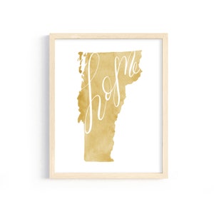 May include: A framed art print featuring the state of Vermont in a gold watercolor design. The word "home" is written in white cursive across the state's outline. The artwork is displayed in a light wood frame.
