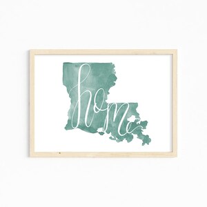May include: A framed watercolor print featuring the state of Louisiana in teal with the word "home" in white script. The artwork is set against a white background, encased in a light wood frame.