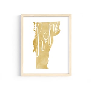 May include: A framed art print featuring the state of Vermont in a golden watercolor design. The word "home" is written in elegant white script across the state's silhouette. The artwork is encased in a light-colored wooden frame.