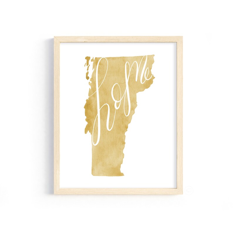 May include: A framed art print featuring the state of Vermont in a golden watercolor design. The word "home" is written in elegant white script across the state's silhouette. The artwork is encased in a light-colored wooden frame.