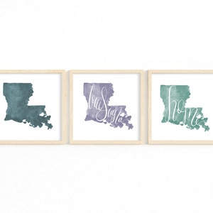 May include: Three framed watercolor prints of the state of Louisiana. Each print features the state's outline in shades of teal, purple, and green. The purple print has the word "Louisiana" in cursive, and the green print has the word "home".