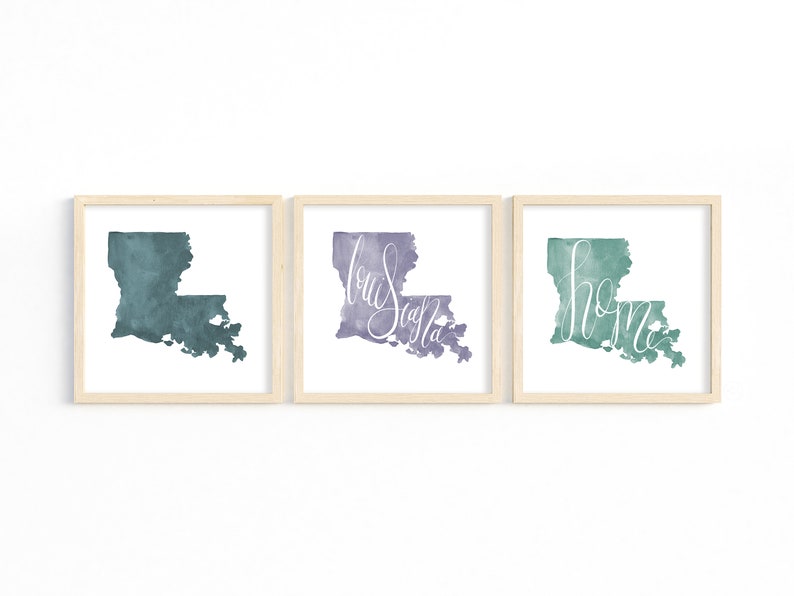 May include: Three framed watercolor prints of the state of Louisiana. Each print features the state's outline in shades of teal, purple, and green. The purple print has the word "Louisiana" in cursive, and the green print has the word "home".