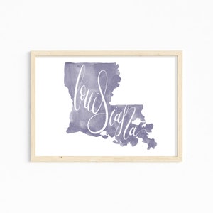 May include: A framed watercolor print featuring the state of Louisiana in a muted purple hue. The word "Louisiana" is written in elegant white script across the state's outline. The frame is made of light-colored wood.