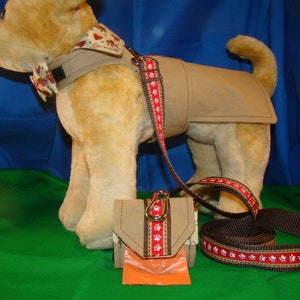 May include: A tan dog coat, harness, leash, and waste bag holder set. The leash and waste bag holder feature a red ribbon with white paw prints. The dog coat has a matching bow tie and neck piece with a heart and paw print pattern.
