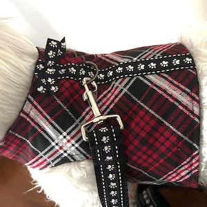 May include: A dog harness featuring a red, black, and white tartan design. The harness is paired with a black lead adorned with white paw prints. A silver metal clasp secures the lead. The harness is trimmed with a black ribbon with white paw prints and hearts.