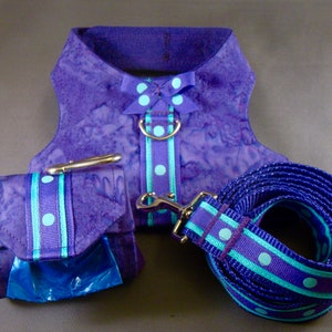 May include: A purple dog harness set with a matching lead and waste bag dispenser. The set features a purple and turquoise polka dot ribbon design. The harness has a bow and a metal ring for lead attachment. The waste bag dispenser includes a blue bag.