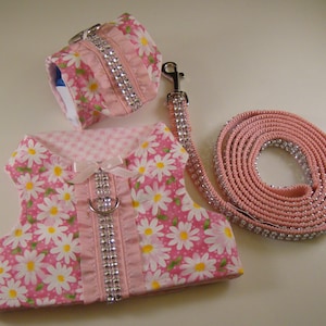 Może przedstawiać: Różowy zestaw dla psa składający się z szelek, obroży i smyczy. Szelki i obroża mają nadruk w stokrotki z białymi kwiatami i żółtymi środkami. Smycz i obroża są ozdobione dżetami.