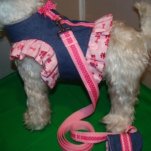 May include: A denim dog harness with a pink polka dot leash and matching accessory. The harness features a pink ruffled skirt with paw and bone prints. A pink bow accents the harness neckline. The leash is approximately 122 cm long.