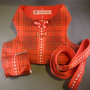 May include: A red plaid dog harness and lead set. The harness features a bow and a gold-coloured ring. The lead and harness have a white paw print design. The set includes a matching waste bag holder. The label reads "Pooch Wear by Penny."