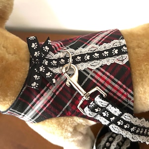 Może przedstawiać: Szelki dla psa z czerwono-czarno-białym wzorem w kratę. Szelki mają czarną wstążkę z białymi odciskami łap i białym koronkowym wykończeniem. Do szelek przymocowany jest srebrny metalowy zatrzask.