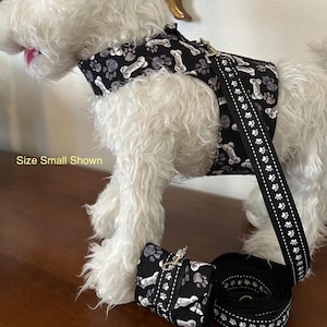 Może przedstawiać: Czarny zestaw dla psa, składający się z uprzęży, smyczy i worka na odpady. Uprząż i worek na odpady mają wzór białych kości i odcisków łap na czarnym tle. Smycz jest czarna z białymi odciskami łap.