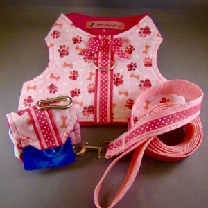 May include: A pink dog harness and lead set with a matching waste bag holder. The harness has a paw print and bone pattern, a pink bow, and a gold-coloured metal ring. The lead and waste bag holder have a polka dot ribbon design.