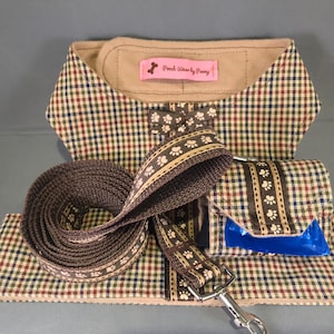 Puede incluir: Un juego de accesorios para perros con un estampado a cuadros marrón, verde y rojo. El juego incluye un collar, una correa, un portabolsas de residuos y una pajarita a juego. La correa y el portabolsas de residuos tienen un diseño de huellas de patas.