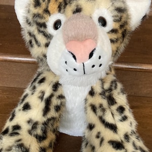 May include: A plush leopard toy with a cream-colored face, ears, and chest. The leopard has black spots on its tan fur, black eyes, and a pink nose. The toy's arms are extended, and the background is a wooden surface.