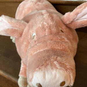 May include: A plush pink pig toy with floppy ears, a rounded snout, and small brown eyes. The stuffed animal is made of soft, fuzzy material and is lying down. The pig has a light pink color.
