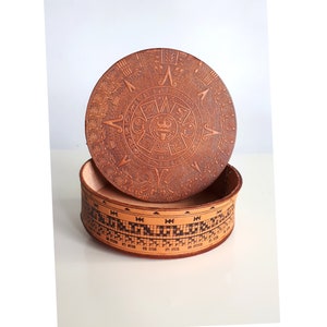 Op de afbeelding: Een ronde, bruine leren doos met een deksel met een ingewikkeld ontwerp van de Azteekse kalender. De doos is versierd met zwarte en bruine geometrische patronen.