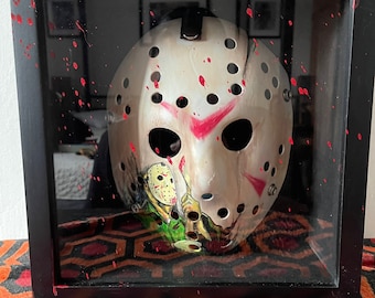 May include: A white hockey mask with red paint splatters and a painted image of a person wearing a hockey mask holding a knife. The mask is displayed in a black frame with a red and black patterned background.