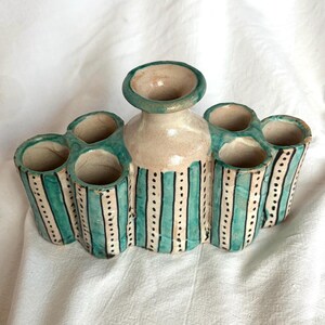 May include: A multi-tube ceramic vase featuring a central neck and six surrounding cylindrical openings. The piece is finished in a cream-colored glaze with vertical teal stripes and thin black dotted lines running down the length of each tube.