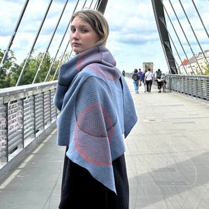 Op de afbeelding: Een blauwe gebreide sjaal met een rood en grijs geometrisch patroon. De sjaal wordt gedragen door een persoon die op een brug staat.