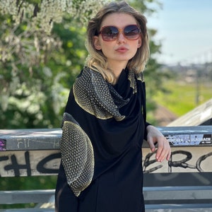 Könnte beinhalten: Eine Frau trägt ein schwarzes Shirt mit Stehkragen und einen gold-schwarzen gemusterten Schal. Sie trägt eine Sonnenbrille und steht auf einem Geländer.