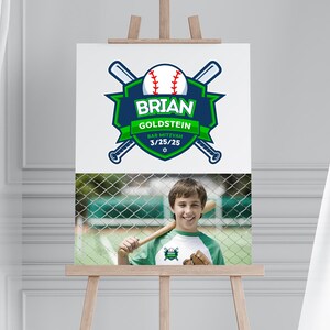 May include: A white sign on an easel features a baseball-themed logo with the name "BRIAN GOLDSTEIN" and "BAR MITZVAH 3/25/25". Below is a photo of a smiling person holding a baseball bat. The logo is green, blue, and white.