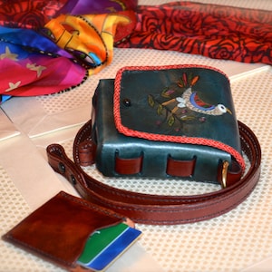 May include: A teal leather purse with a red trim and a bird design. The purse has a brown leather strap and a small brown leather wallet attached to the strap.