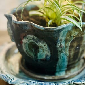 May include: A handmade ceramic planter with a wavy, scalloped rim and a matching saucer. The glaze features a deep, dark blue and black base with swirling teal and turquoise accents. A small green and white spider plant grows inside the pot.