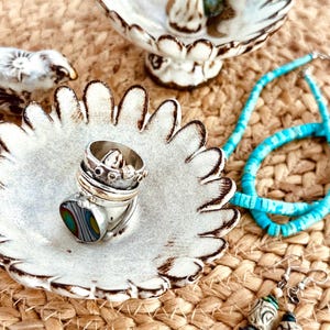 May include: Two white ceramic dishes with scalloped edges, one holding a stack of silver rings with a colorful inlay. A turquoise beaded necklace and a pair of dangling earrings with patterned beads are arranged alongside the ceramic dishes.