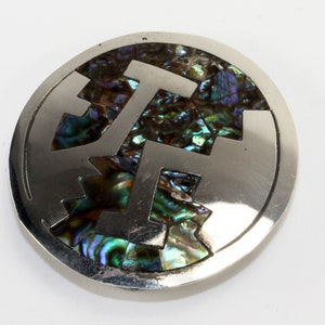 May include: A silver circular brooch with a geometric design inlaid with iridescent abalone shell. The shell is a mix of green, blue, and brown.