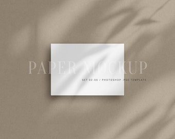 May include: A white card mockup with a shadow of leaves on it. The card is on a beige background. The text "PAPER MOCKUP SET 02-06 / PHOTOSHOP PSD TEMPLATE" is printed on the card.