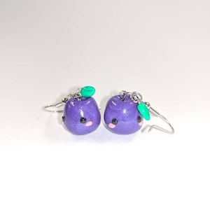 May include: A pair of purple dangle earrings with a green leaf charm and a cute face. The earrings are made of polymer clay and have a sparkly finish.
