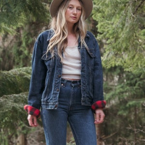 May include: A woman wearing a denim jacket with red and black plaid lining, blue jeans, and a beige hat. She is standing in a forest setting.