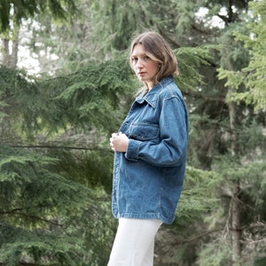May include: A woman wearing a blue denim jacket and white wide-leg pants stands on a wooden deck in front of a green forest.