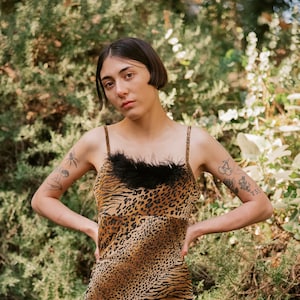 May include: A woman wearing a short, brown and black animal print dress with a black feather trim at the neckline. The dress has a fitted silhouette and a straight hemline. The woman is standing in front of a green leafy background.